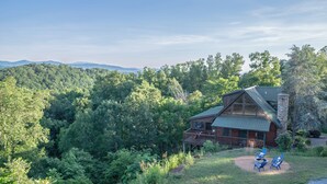 Une cabane dotée d’un porche et d’une terrasse, entourée d’une forêt dense et avec des montagnes en arrière-plan.