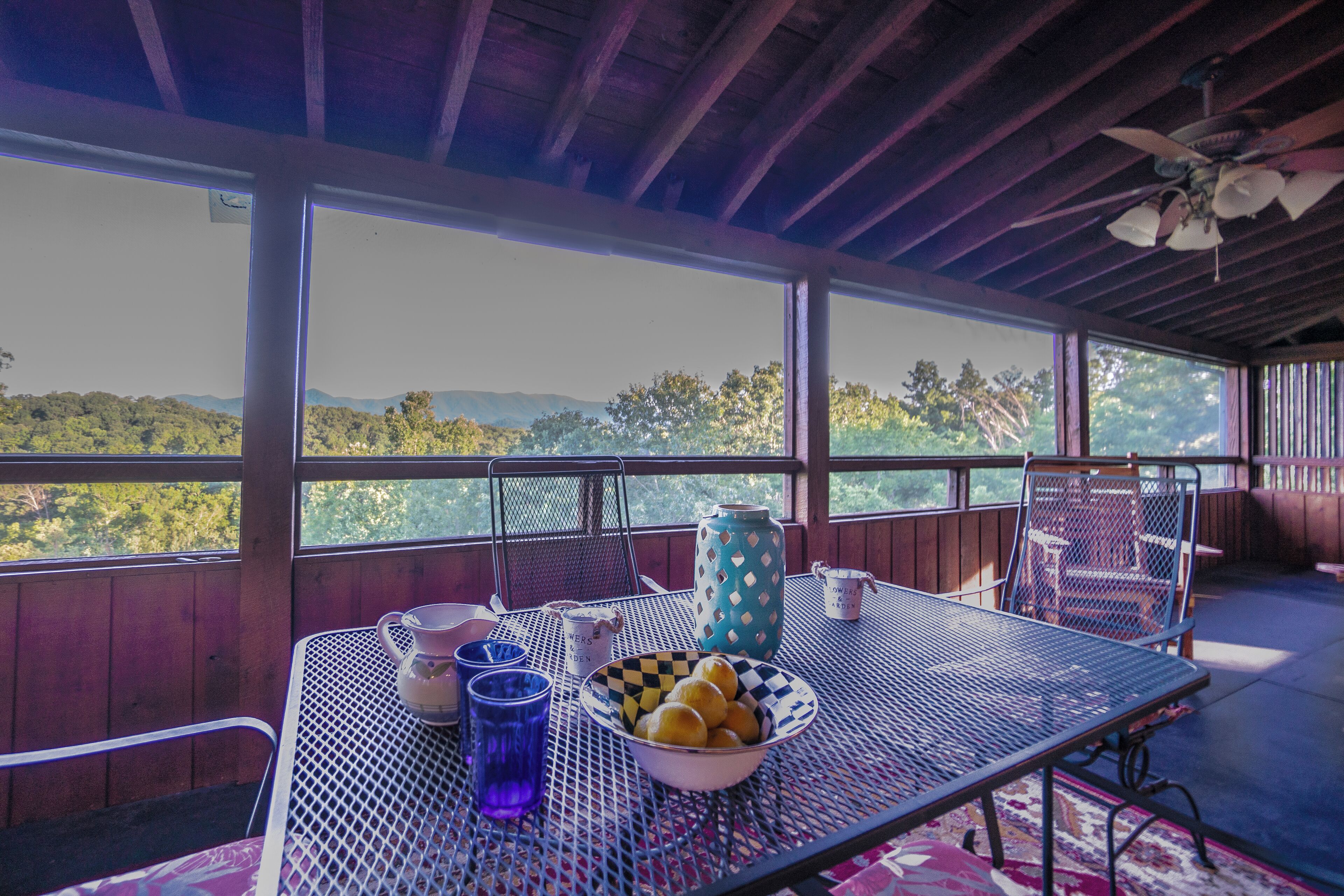 Un patio techado con una mesa lista para comer, ventanas amplias que ofrecen vistas del paisaje y un ventilador de techo.