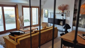 Un salon moderne avec un canapé jaune, une table à manger et une vue sur l’extérieur grâce à de grandes fenêtres.