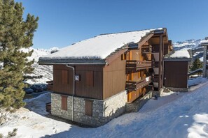A wooden chalet with a stone foundation, a wooden staircase, and a snow-covered roof, surrounded by mountains.