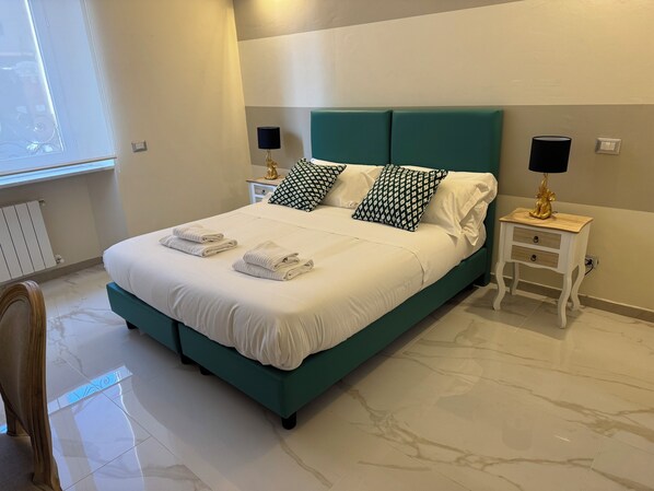 Una cama bien hecha con sábanas blancas y dos almohadones estampados, flanqueada por mesitas de noche con lámparas, en una habitación con piso de mármol y una ventana con persianas.