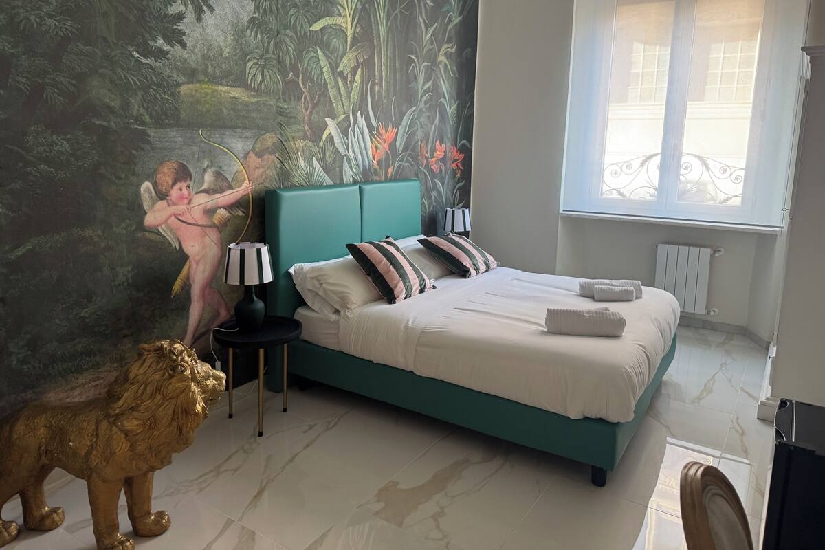Une chambre à coucher avec un lit de couleur turquoise, du linge de lit blanc, une fresque représentant une scène de forêt, une statue de lion dorée et une fenêtre donnant sur un balcon.