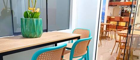 A dining area with a wooden table and turquoise chairs, a green vase with white flowers, and a view into a restaurant through a glass partition.