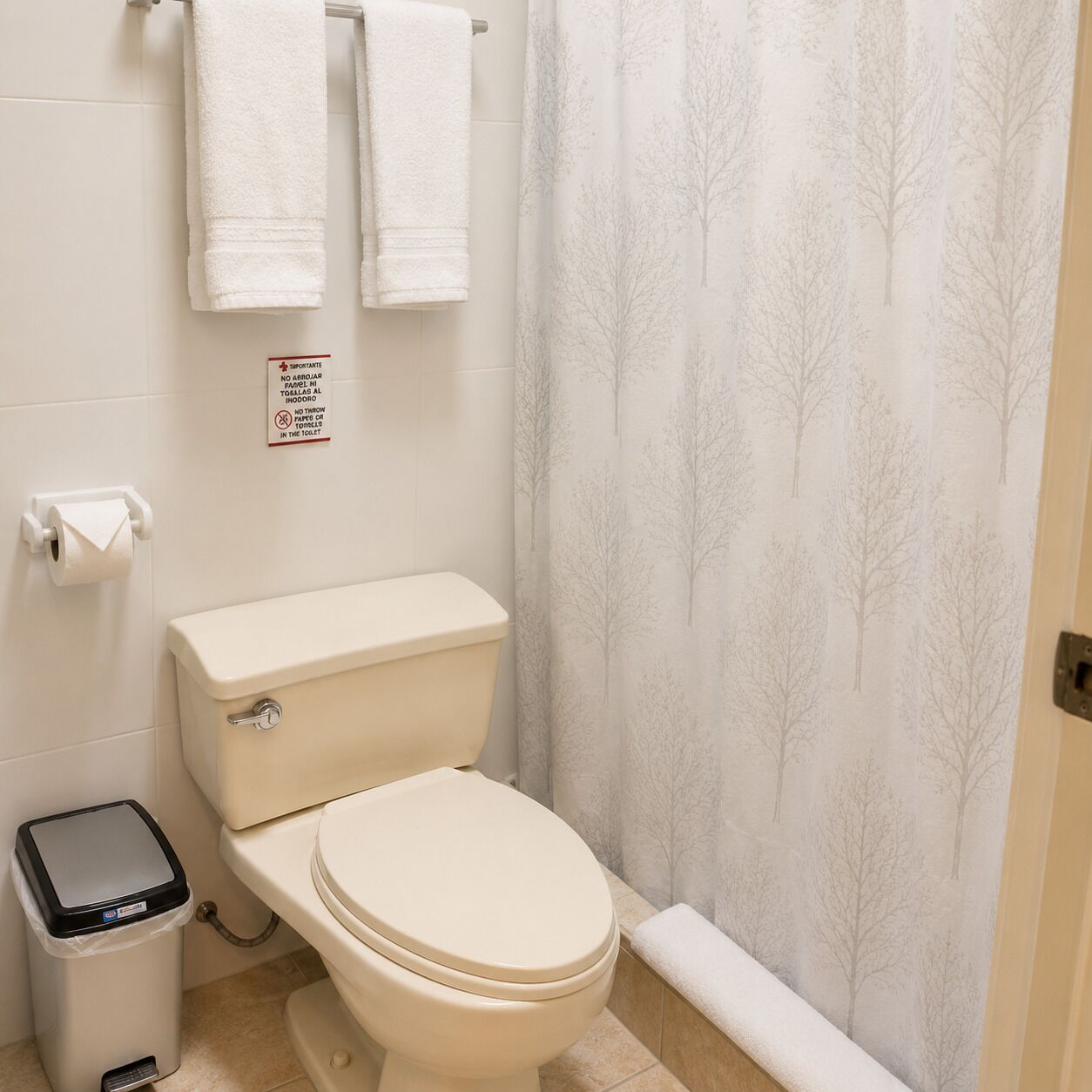 A bathroom with a toilet, a trash bin, and a shower curtain with a tree pattern.