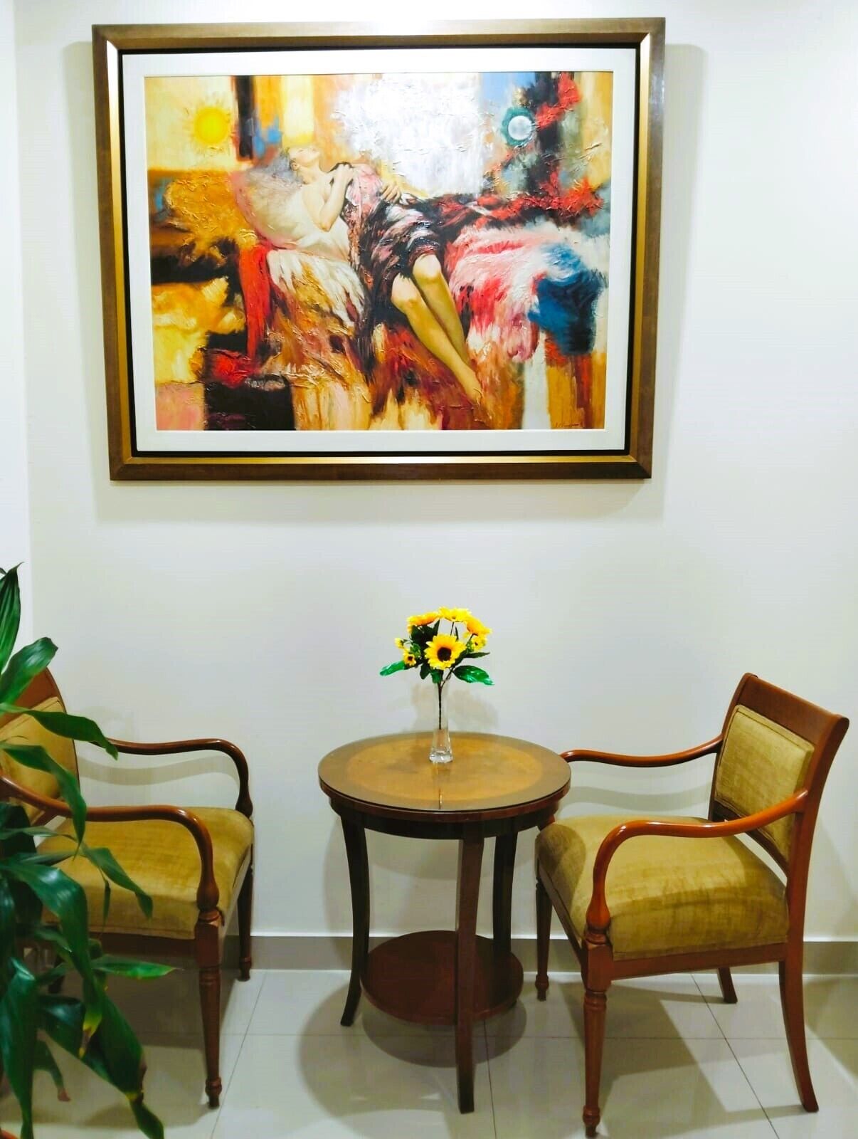 A room with two wooden chairs, a small round table with a vase of sunflowers, and a large abstract painting on the wall.