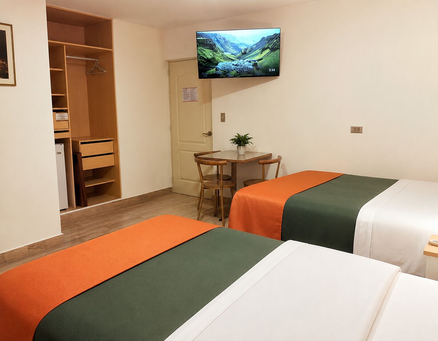 Habitación de hotel con dos camas, un escritorio con silla, televisor montado en la pared y un armario.