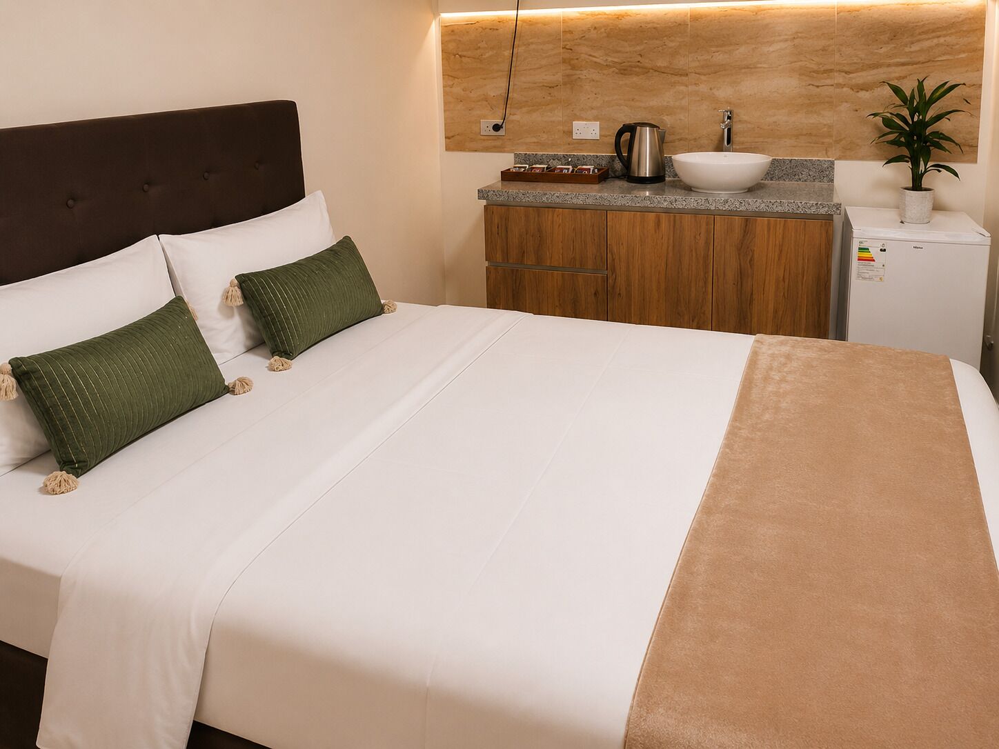 Habitación de hotel con cama, una kitchenette pequeña, una planta y una lámpara fijada a la pared.