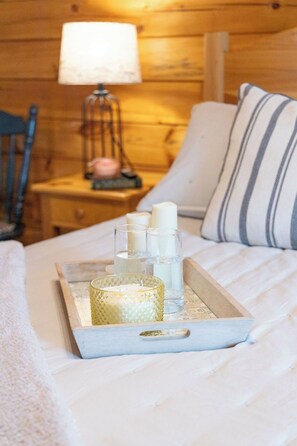 Interior de una cabaña de madera con una cama, una mesita de noche, una lámpara y una bandeja con velas y candelabro.