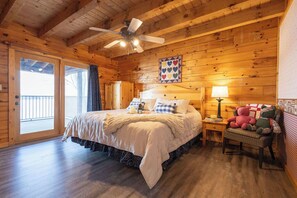 A cozy wooden cabin bedroom with a large bed, a ceiling fan, a nightstand with a lamp, and a chair with stuffed animals.