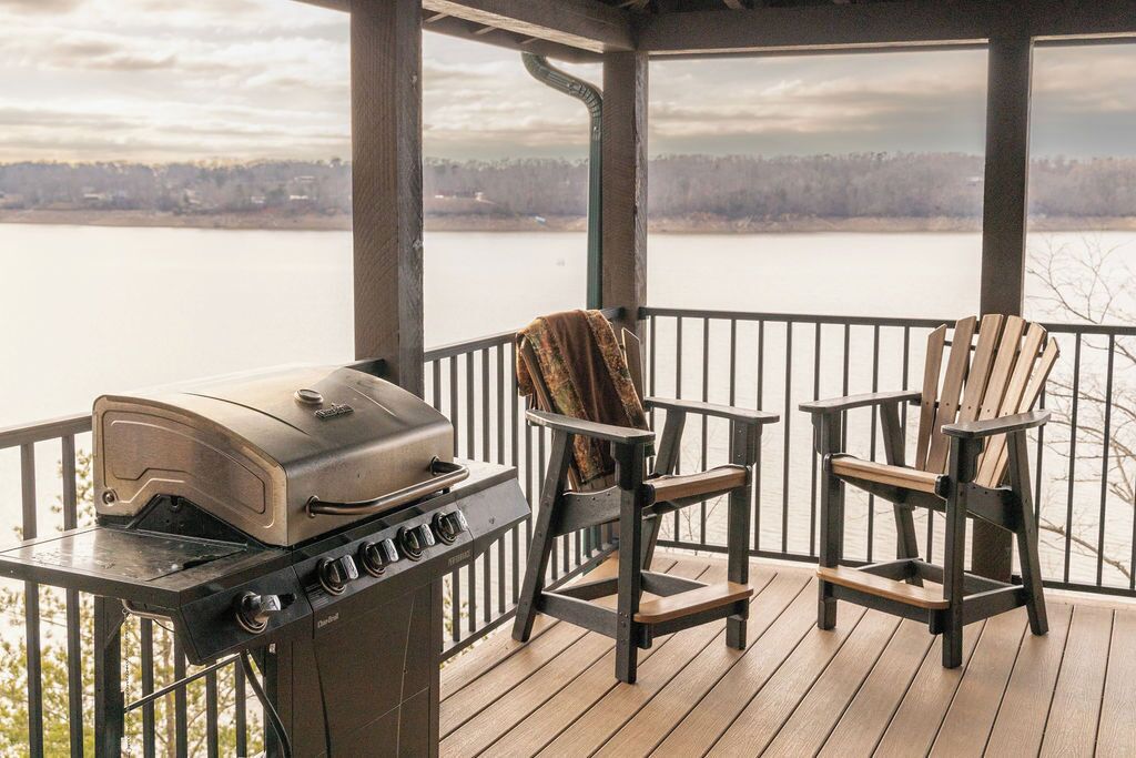 A covered outdoor area with a grill, two Adirondack chairs, and a view of a lake.