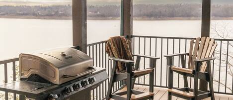 A covered outdoor area with a grill, two Adirondack chairs, and a view of a lake.