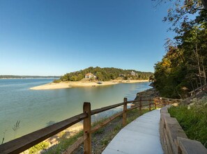 A lakeside property with a wooden deck and a stone wall, overlooking a calm lake and a forested area with houses.
