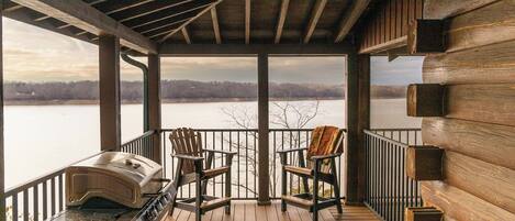 A wooden deck with a grill and two Adirondack chairs overlooking a lake.