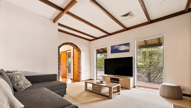 A modern living room with a grey sofa, a wooden coffee table, and a flat-screen TV. There is a large window with blinds, a ceiling fan, and a wooden ceiling with exposed beams.