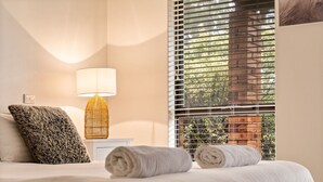 A neatly made bed with two rolled towels, a fluffy pillow, a bedside lamp, and a window with blinds.