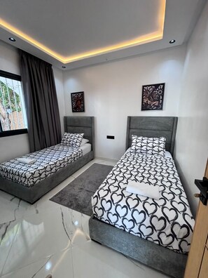 Quarto moderno com duas camas de solteiro, uma janela grande com cortinas e iluminação embutida no teto.