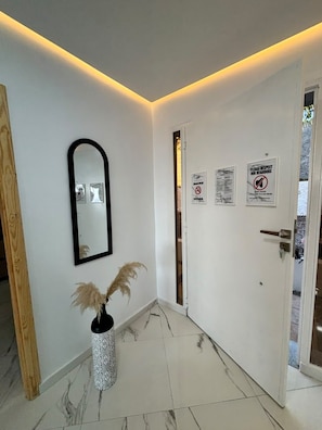 A hallway with a white door, a mirror, and a vase with dried plants. There are signs on the door indicating no smoking and respect for neighbors.