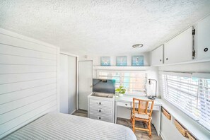 A bedroom with a bed, a desk, and a chair. There is a window with blinds, a door, and wall-mounted artwork.