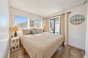 Un dormitorio con una cama grande, una mesita de noche con lámpara, un reloj en la pared y vistas al exterior a través de una ventana.