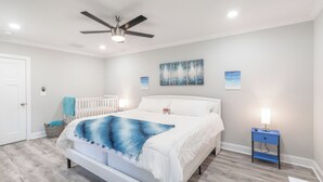 Une chambre à coucher avec un lit blanc, un ventilateur de plafond, un berceau blanc et une table de chevet bleue avec une lampe.