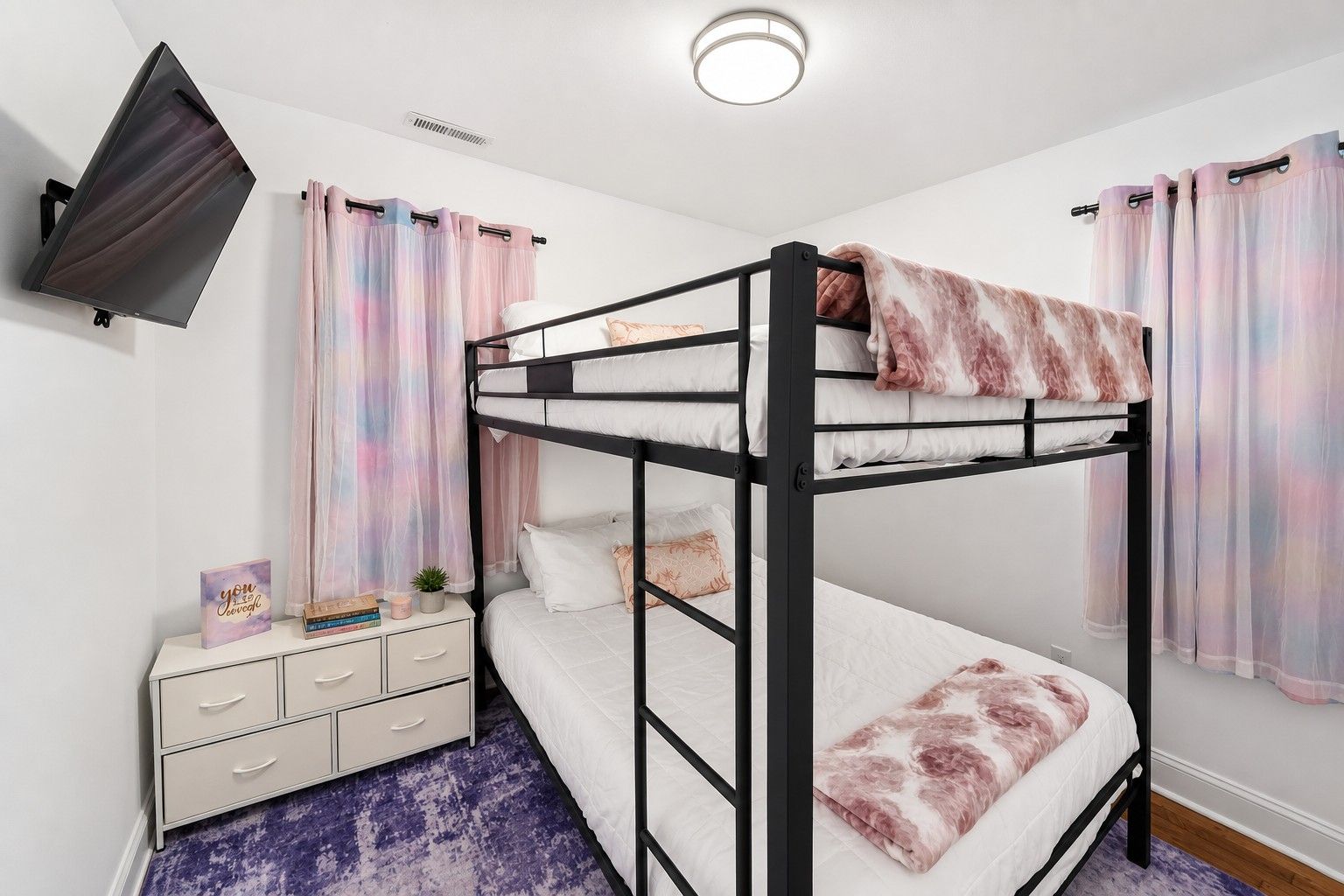 Habitación con literas, televisor montado en la pared, mesita de noche con cajones y alfombra púrpura.