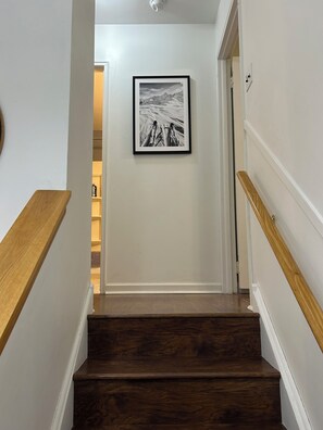 Un escalier en bois, avec des murs blancs et un tableau encadré représentant un paysage de montagne.