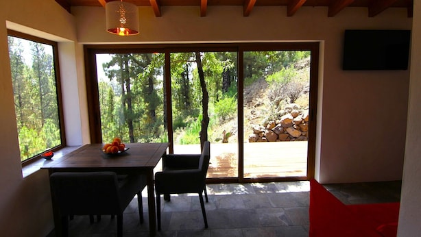 Un coin repas avec une table et des chaises, un saladier de fruits, et une vue sur un paysage naturel grâce à de grandes portes-fenêtres coulissantes.