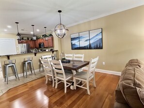 A dining area with a wooden table and white chairs, a kitchen with wooden cabinets, and a painting on the wall.