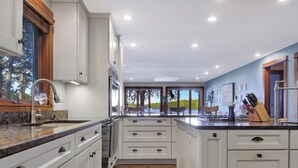 A modern kitchen with white cabinetry, a granite countertop, and a view of a lake through large windows. - Spacious 5-bedroom lakefront home on pristine Lower Narrows Pond in Winthrop (Winthrop)
