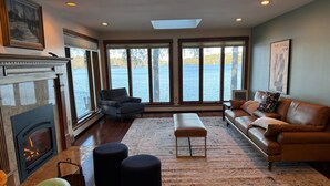 A spacious living room with a fireplace, large windows overlooking a body of water, a leather sofa, and a modern design. - Spacious 5-bedroom lakefront home on pristine Lower Narrows Pond in Winthrop (Winthrop)