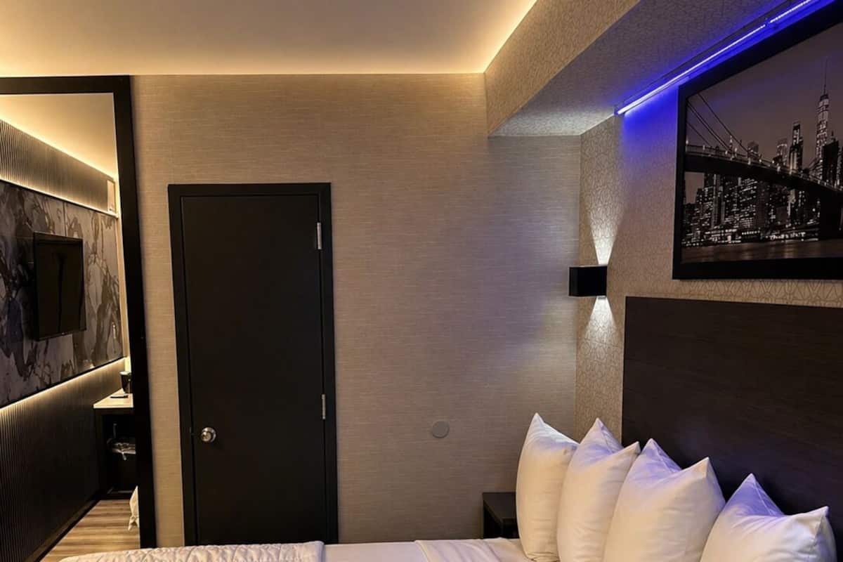 A modern hotel room with a large bed, a dark door, a wall-mounted TV, and a cityscape picture.