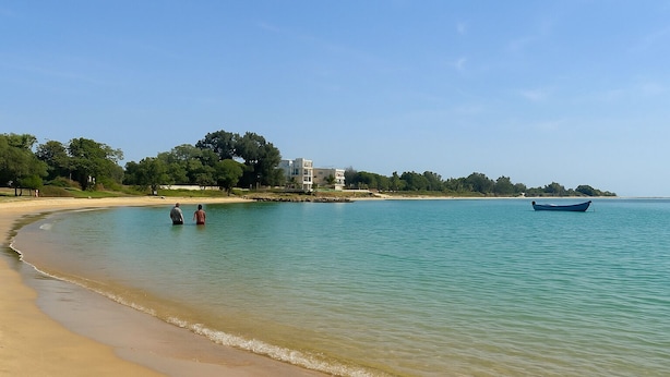 Una playa con agua cristalina, un bote y un edificio al fondo.