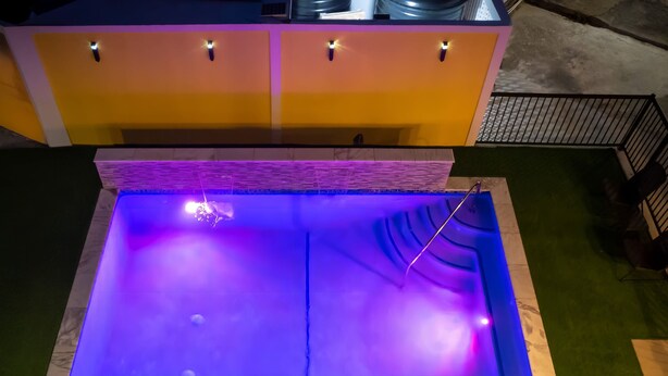 Piscina rectangular con iluminación azul en su interior, rodeada por un césped verde y un edificio amarillo con base de ladrillo.