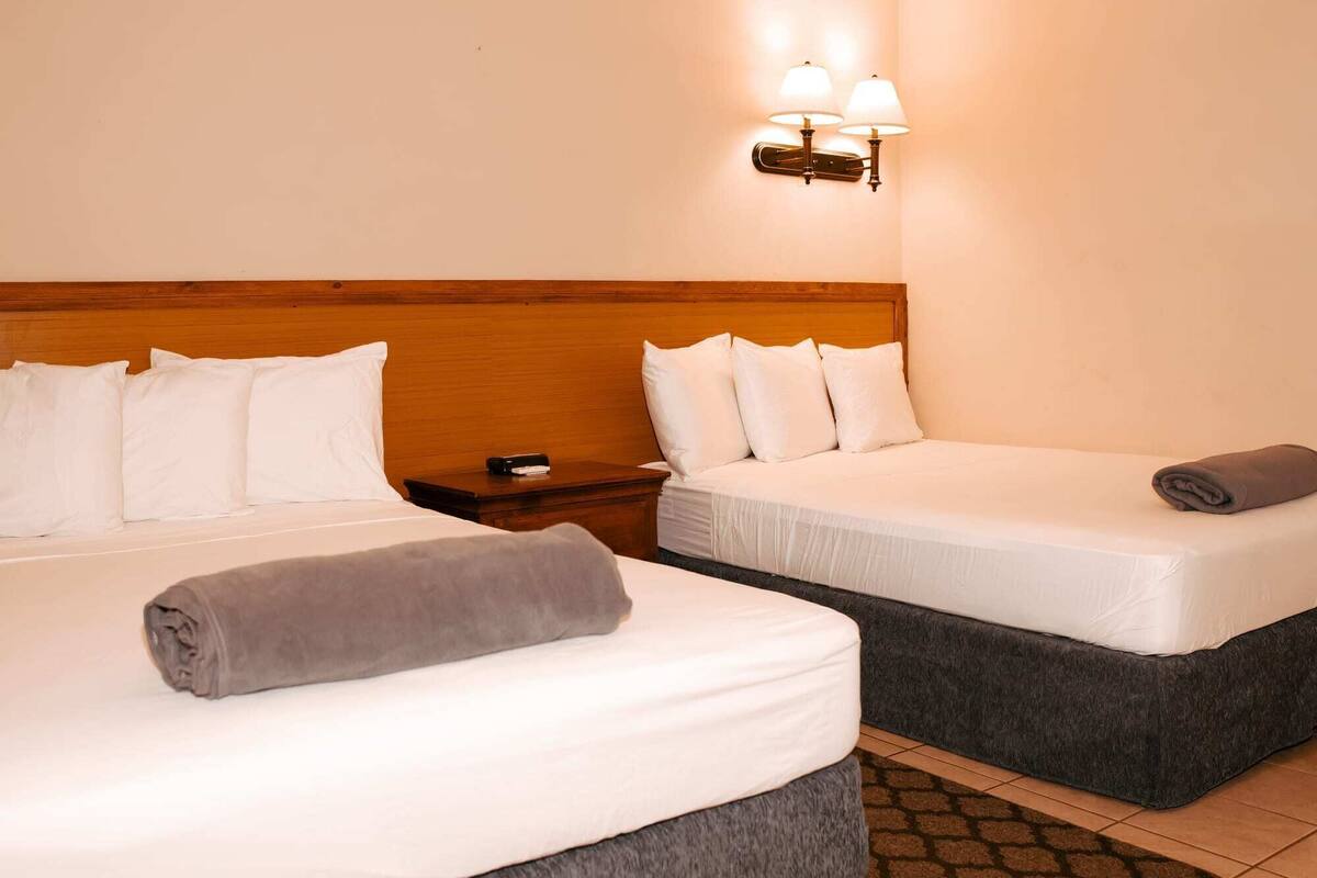 A hotel room with two beds, a wooden headboard, white bedding, a nightstand with a lamp, and a patterned rug.