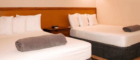 Habitación de hotel con dos camas, cabecera de madera, ropa de cama blanca, mesita de noche con lámpara y alfombra estampada.