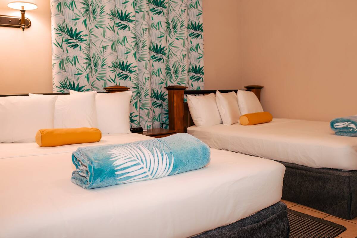 A hotel room with two beds, each with white bedding and a single pillow. There is a wooden nightstand between the beds, and a lamp on the wall.