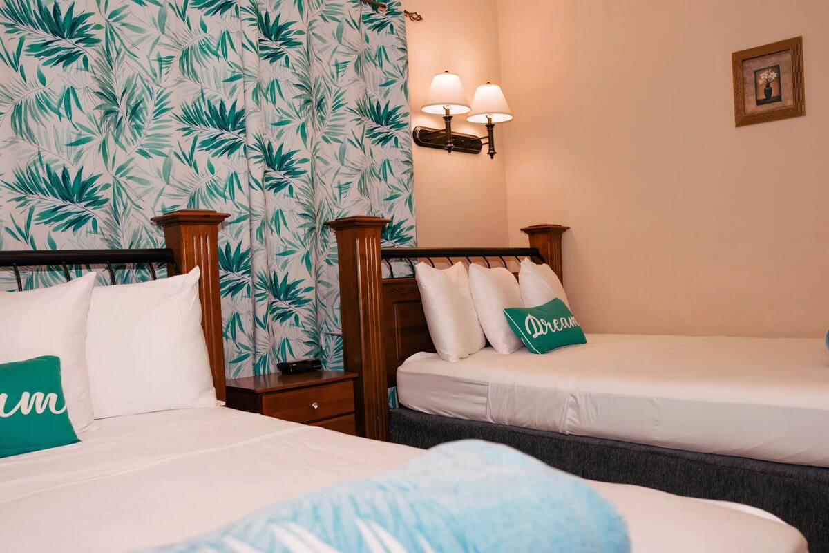 A hotel room with two beds, a nightstand, a lamp, and a wall with a tropical leaf pattern.