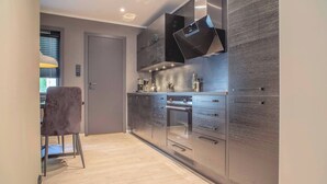 A modern kitchen with stainless steel appliances, a grey upholstered chair, and a door leading to another room. - Cozy Ålesund Apartment With Private Entrance & Free Parking (Ålesund)