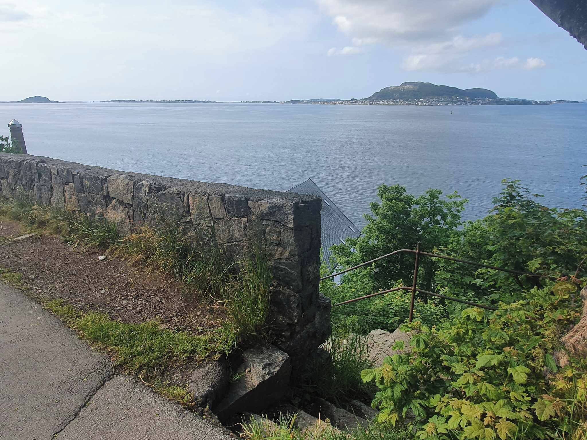 Vista costera con muro de piedra, zona verde y una imagen clara del mar y la costa distante.