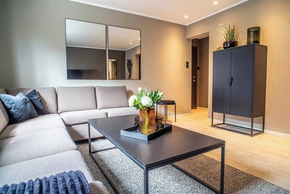 A modern living room with a sectional sofa, a coffee table with a vase of flowers, a large mirror, and a cabinet. - Cozy Ålesund Apartment With Private Entrance & Free Parking (Ålesund)