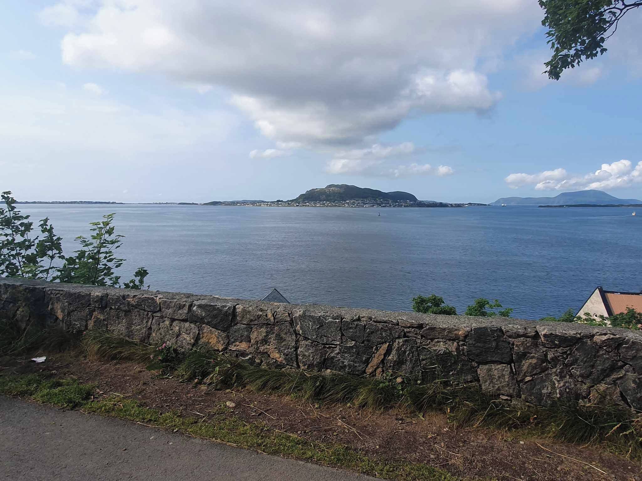 Blick auf die Küste mit einer Steinmauer im Vordergrund, einem Gewässer und weit entfernten Landformen unter einem teilweise bewölkten Himmel.