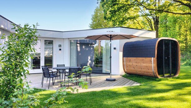 Eine moderne Terrassenanlage mit Essplatz, großem Sonnenschirm und einer zylindrischen Struktur – vermutlich eine Sauna oder ein Whirlpool – inmitten eines üppigen Gartens.