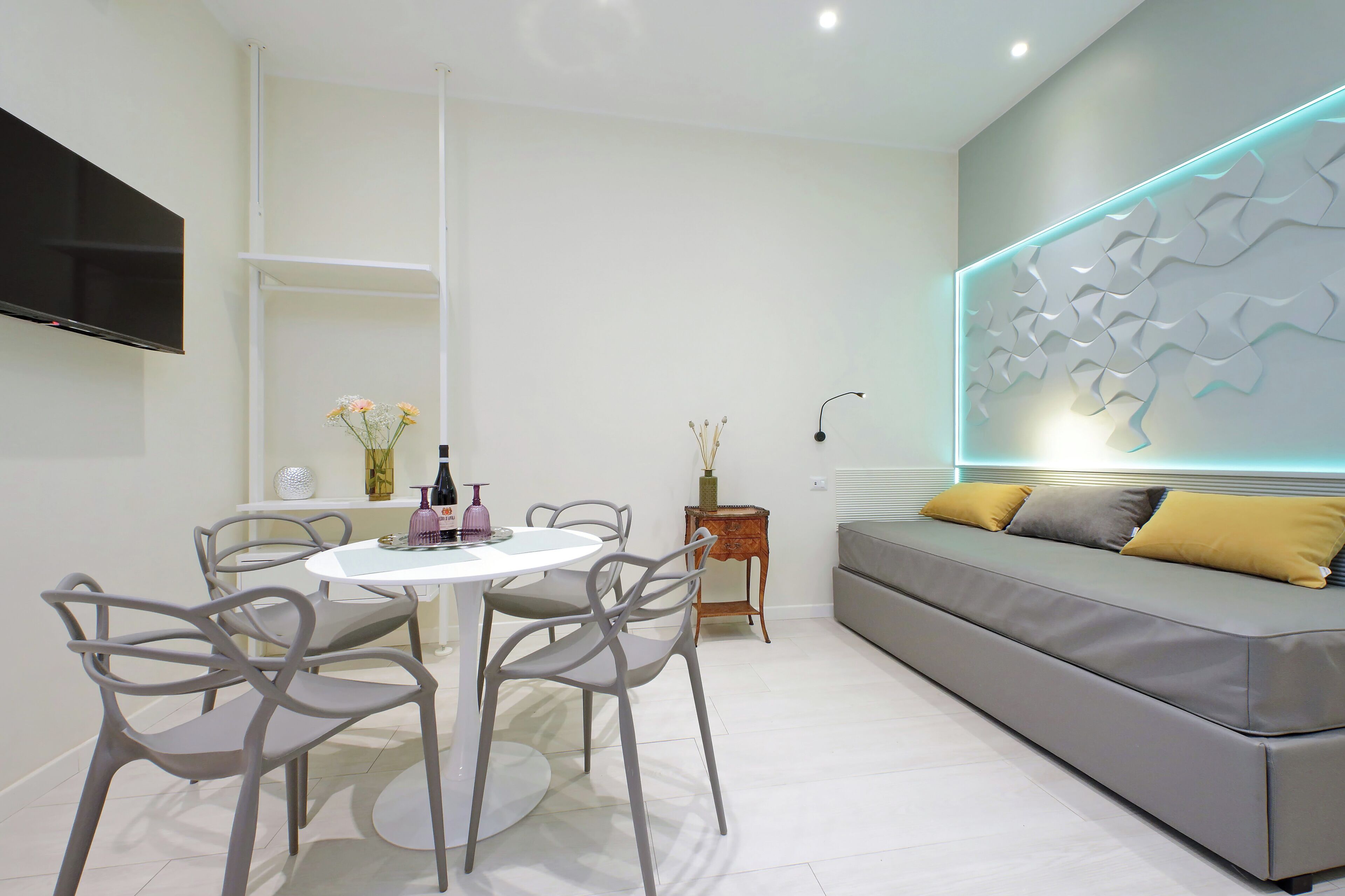 A modern living room with a grey sofa, a white dining table with four chairs, and a flat-screen TV mounted on the wall.