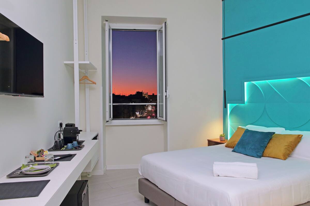 A hotel room with a bed, a desk with a coffee machine, a TV, and a window with a city view.