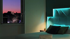 A hotel room with a bed, a nightstand, a window with a city view, and a headboard with built-in lighting. - Domus Augustea San Giovanni Sunset Suites (Rome)