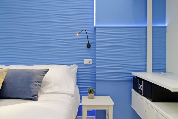 A bedroom with a bed, a nightstand, a small plant, and a wall-mounted light fixture. - Domus Augustea San Giovanni Sunset Suites (Rome)
