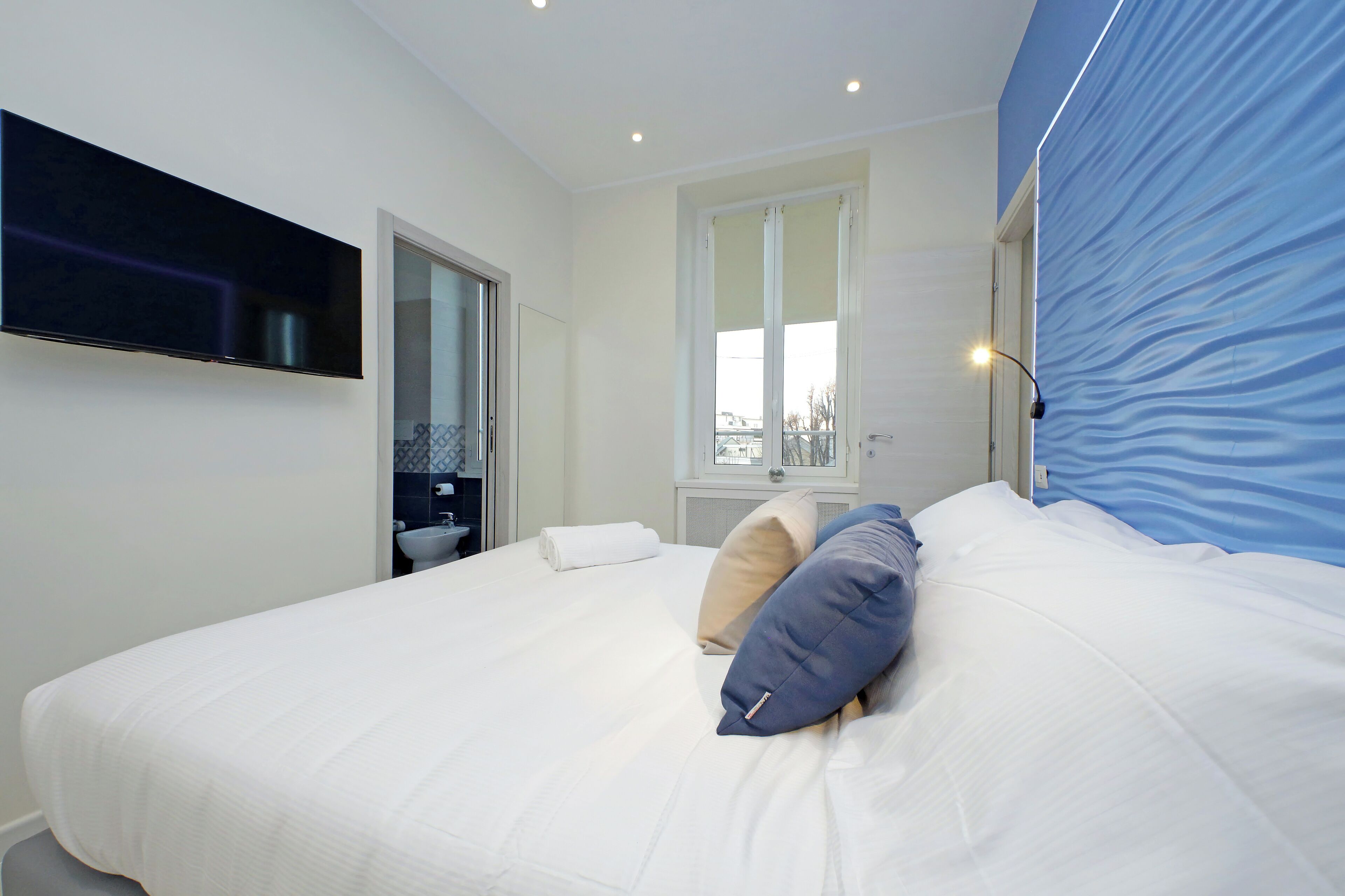 A hotel room with a large bed, a flat-screen TV mounted on the wall, a bathroom with a sink and mirror, and a window with a view of the outside.
