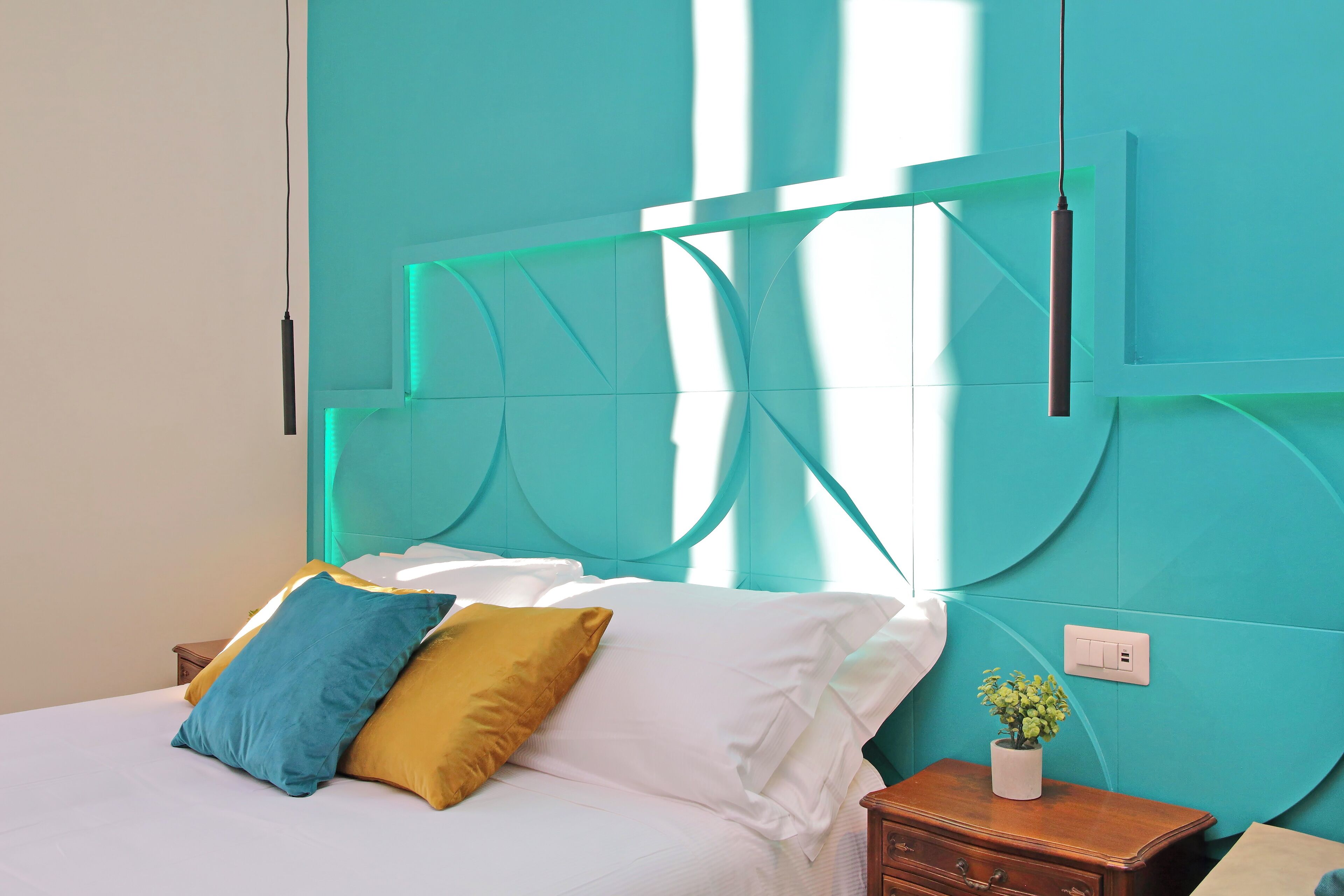 Uma cama com almofadas brancas e azuis, uma cabeceira verde-água e uma pequena mesa de cabeceira de madeira com uma planta.