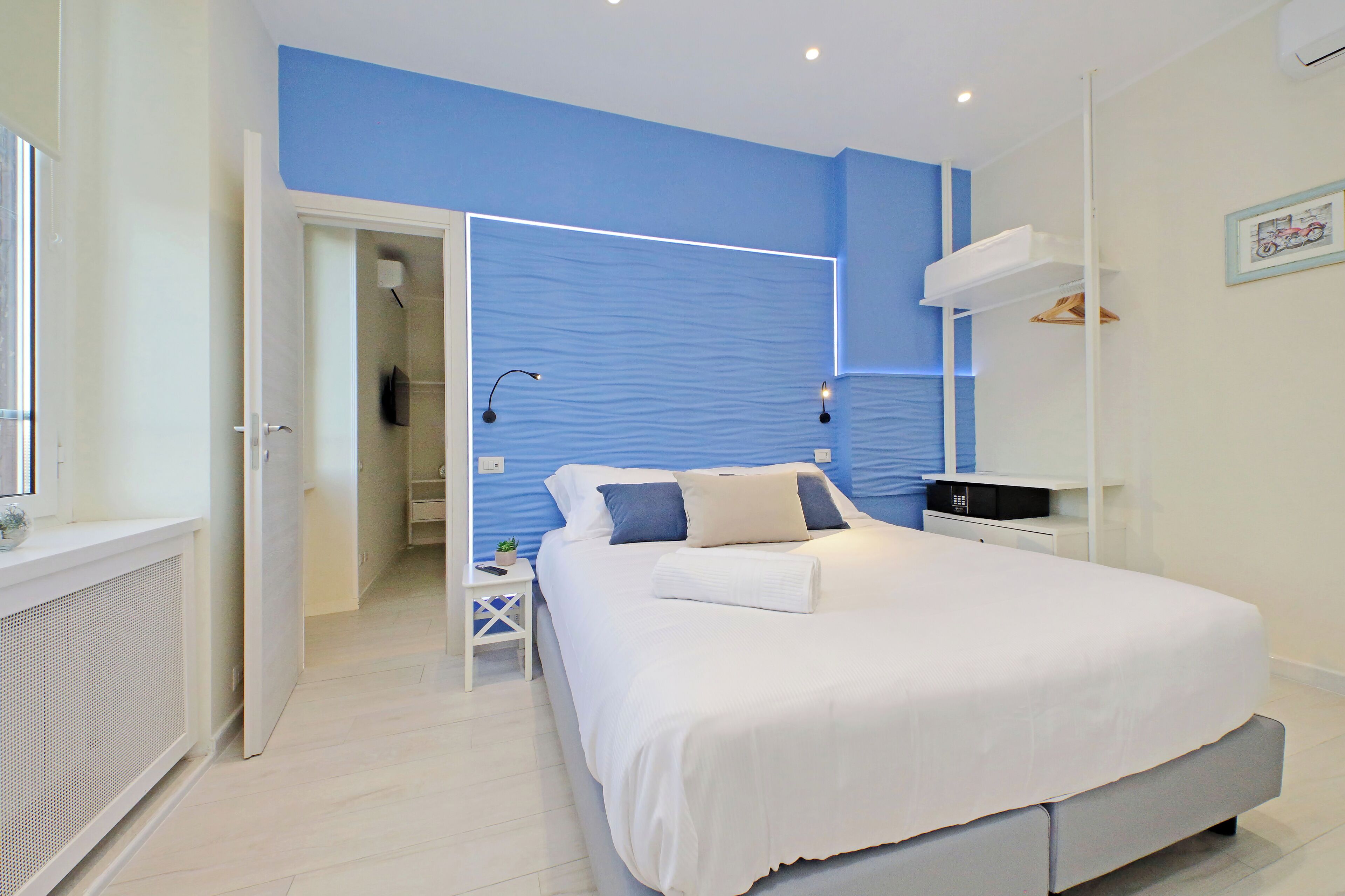 A bedroom with a large bed, a bedside table, a nightstand, a wall-mounted TV, and a door leading to another room.