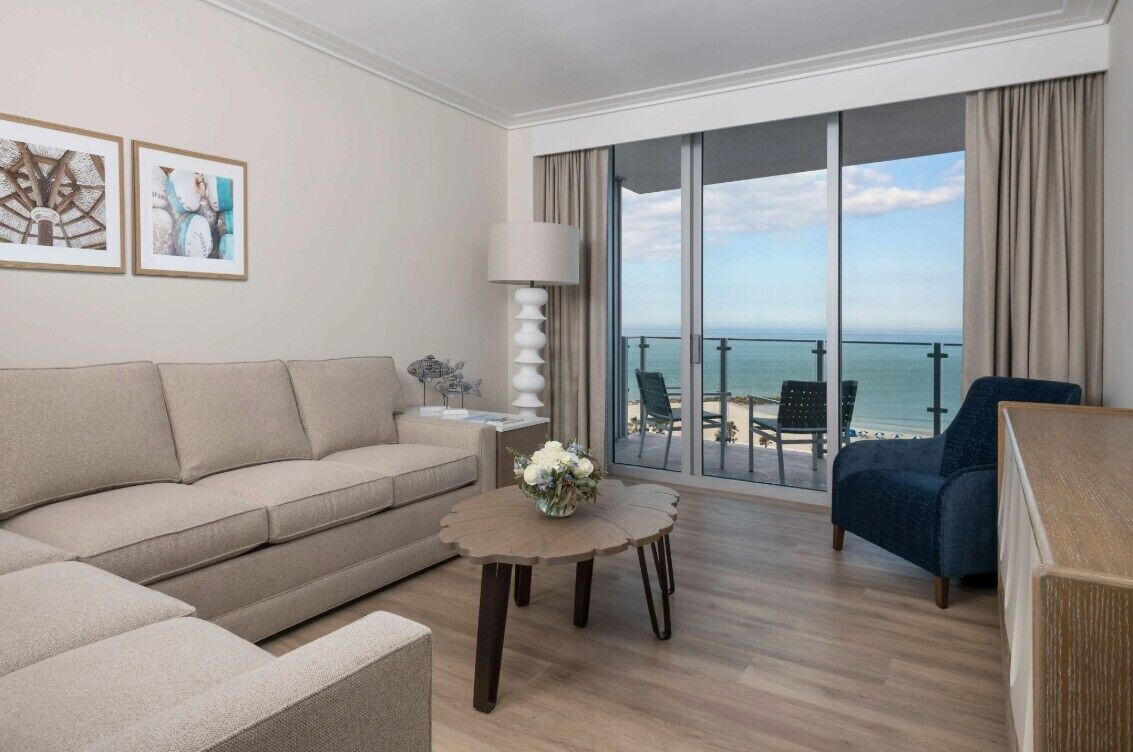 A modern hotel room with a large balcony, a sofa, a round coffee table, and a chair.